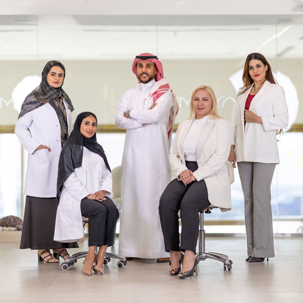 A team section for MAYAM Aesthetics, showcasing a group photo of diverse professionals, including men and women dressed in white coats and business attire, posing in a modern clinic setting. The text highlights their expertise in aesthetics and wellness, emphasizing their commitment to delivering high-quality care. A button labeled 'Explore Our Team of Experts' invites users to learn more about the specialists.