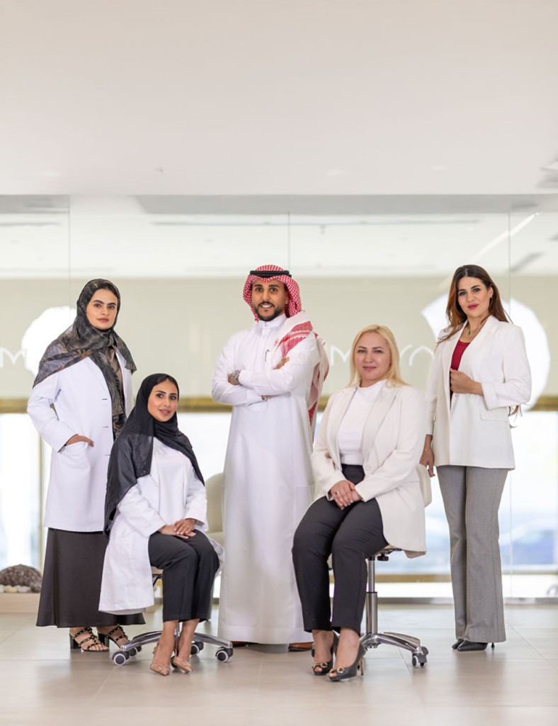 A team section for MAYAM Aesthetics, showcasing a group photo of diverse professionals, including men and women dressed in white coats and business attire, posing in a modern clinic setting. The text highlights their expertise in aesthetics and wellness, emphasizing their commitment to delivering high-quality care. A button labeled 'Explore Our Team of Experts' invites users to learn more about the specialists.