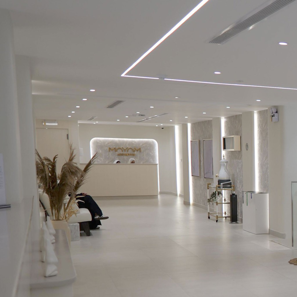A modern and elegant reception area of MAYAM Aesthetics clinic, featuring a minimalist design with white walls, LED ceiling lighting, decorative plants, and a reception desk in the background. The image is part of the 'Contact Us' section on their website.