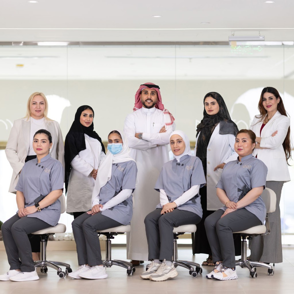 A group photo of a professional medical or aesthetic team from MAYAM Aesthetics, featuring a diverse staff including men and women in white coats and gray uniforms, posing in a modern clinic setting. The image is part of a Frequently Asked Questions (FAQs) section on their website.