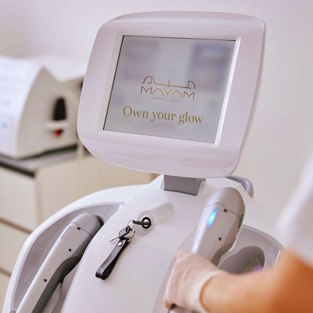 Advanced aesthetic treatment machine at Mayam Aesthetics clinic with a glowing screen displaying 'Own your glow' message.